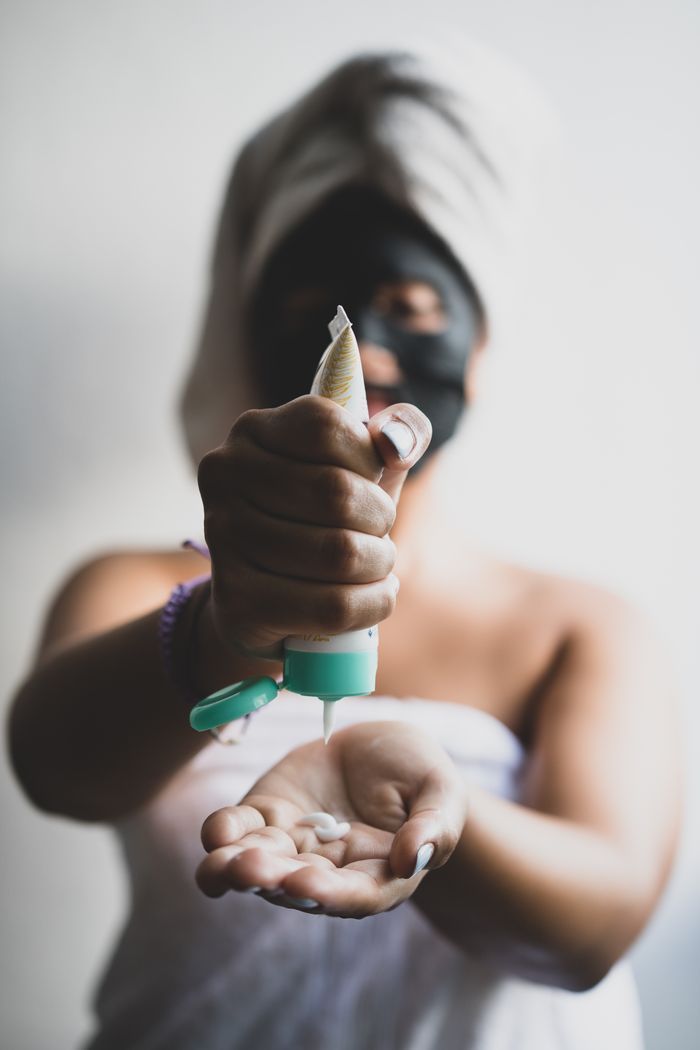 about-us Close-up of a woman applying facial cream with a face mask and towel headwrap at home.